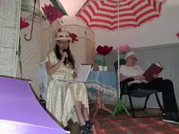 Kobieta w sukni i kapeluszu recytuje, mężczyzna w czyta w fotelu. W tle parasole i kwiaty.