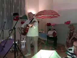 Mężczyzna gra na gitarze, a w tle siedzą osoby w kapeluszach. Kolorowe parasole dekorują przestrzeń.