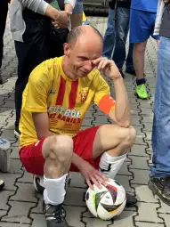 Mężczyzna w żółto-czerwonym stroju piłkarskim siedzi na ziemi z piłką obok.