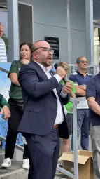 Mężczyzna w garniturze przemawia z pasją, w tle grupa osób słuchających.