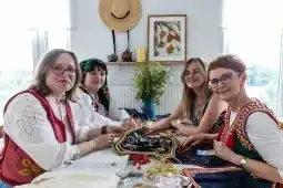 Cztery kobiety w tradycyjnych strojach haftują i rozmawiają w jasnym wnętrzu.