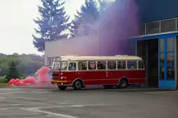 Czerwony autobus wyjeżdża z budynku, otoczony kolorowym dymem.