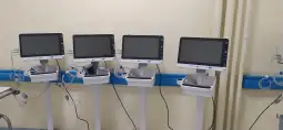 Cztery monitory medyczne na stojakach w szpitalnym pomieszczeniu.