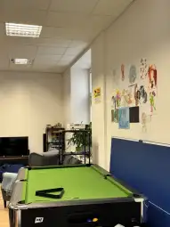 Stół bilardowy z zielonym suknem w biurze, na ścianie rysunki i rośliny w tle.
