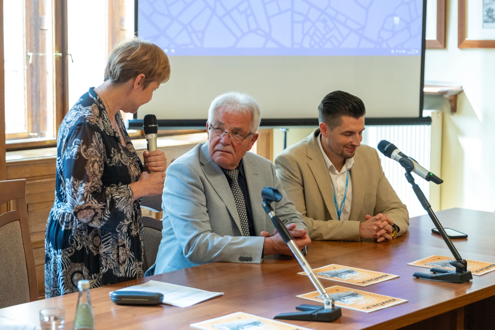Spotkanie trzech osób przy stole, jedna z nich mówi do mikrofonu, w tle ekran.  Autor: Bogusław Świerzowski