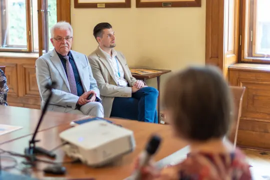 Dwóch mężczyzn słuchających prezentacji w sali konferencyjnej.