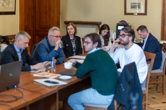 Grupa ludzi siedzących przy stole podczas spotkania, z laptopami i notatkami.
