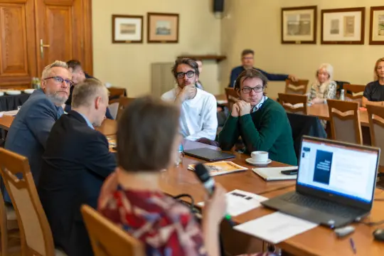 Grupa ludzi uczestniczy w dyskusji przy stole w sali konferencyjnej.