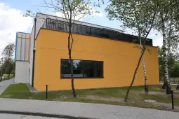 Nowoczesny budynek z żółtą elewacją i dużym oknem, otoczony drzewami.