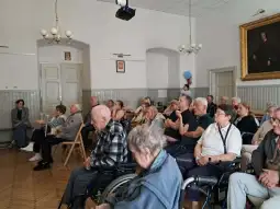Grupa starszych osób siedzących w sali, słuchających wykładu lub prezentacji.