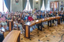 Grupa młodzieży siedząca w sali obrad, z zainteresowaniem słuchająca prelekcji.