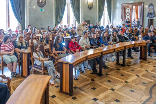 Grupa młodzieży siedząca w sali obrad, z zainteresowaniem słuchająca prelekcji.