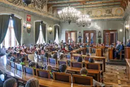 Sala obrad z dużą grupą ludzi, w tym dzieci, siedzących w rzędach.