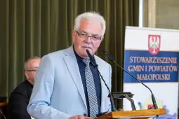 Mężczyzna w jasnym garniturze przemawia do mikrofonu na tle banneru Stowarzyszenia Gmin i Powiatów Małopolski.