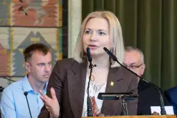 Kobieta przemawia do zgromadzenia, w tle mężczyźni słuchają.