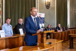 Mężczyzna w garniturze przemawia przy mównicy w sali konferencyjnej. W tle siedzą inni uczestnicy.