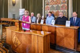 Kobieta przemawia przy mównicy, obok siedzi grupa osób w sali konferencyjnej.