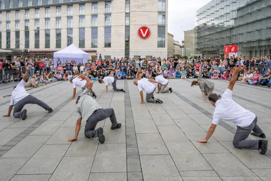 Grupa tancerzy wykonujących układ taneczny na placu, otoczona widownią.