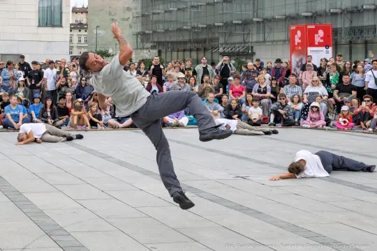 Tancerz w skoku na tle publiczności, inni tancerze leżą na ziemi.