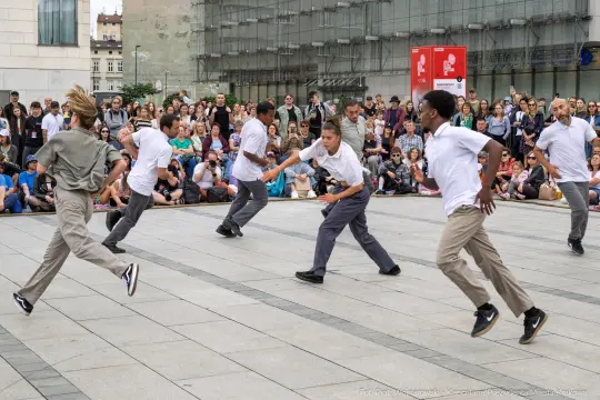 Grupa tancerzy wykonujących choreografię na placu, otoczona widownią.