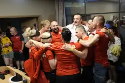 Grupa ludzi w czerwonych koszulkach świętuje zwycięstwo, uśmiechając się i przytulając.