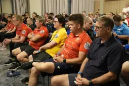 Grupa ludzi siedzących w sali, ubranych w koszulki sportowe, skupionych na prezentacji.