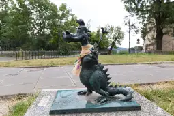Rzeźba smoka w szaliku, stojąca na chodniku w otoczeniu zieleni.