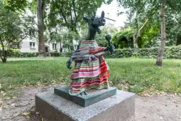Rzeźba psa w kolorowej sukience, trzymającego kwiat, w parku otoczonym drzewami.