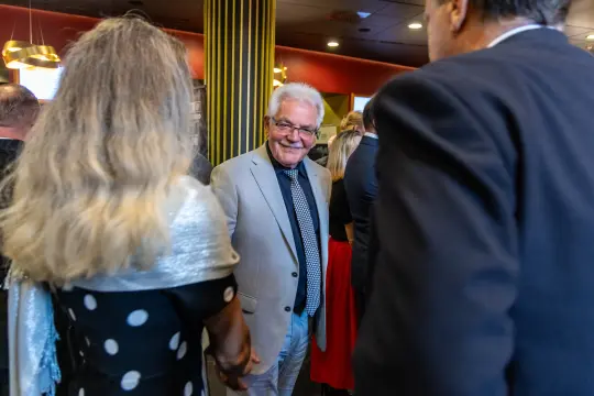 Mężczyzna w jasnym garniturze uśmiecha się do osób w eleganckich strojach.