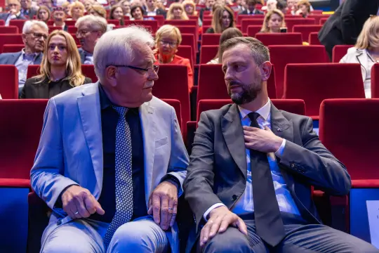 Dwaj mężczyźni w garniturach rozmawiają na tle widowni w sali.