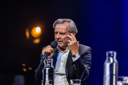 Mężczyzna w garniturze trzyma mikrofon i gestykuluje podczas wystąpienia.
