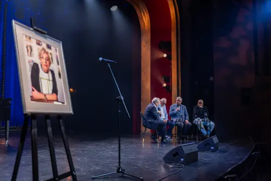 Panel dyskusyjny na scenie z portretem osoby na sztaludze.