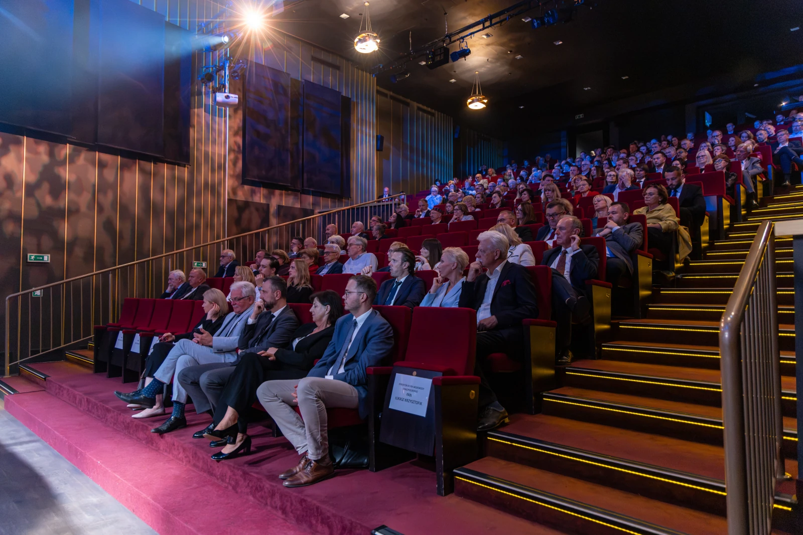 Widownia w teatrze z siedzącymi widzami, oświetlenie sceny i czerwone fotele.  Autor: Bogusław Świerzowski