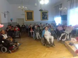 Grupa osób starszych w wózkach inwalidzkich w sali z dekoracjami i obrazami na ścianach.