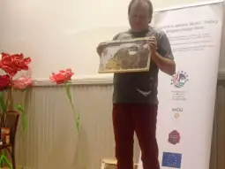 Mężczyzna trzyma ramkę z plastrami miodu podczas prezentacji. W tle kwiaty i baner promocyjny.