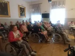 Grupa osób starszych na wózkach inwalidzkich w sali, uczestniczących w spotkaniu.