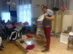 Mężczyzna prezentuje książkę przed grupą osób w sali z dekoracjami i kwiatami.