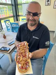 Mężczyzna trzyma długi hot dog z ketchupem i posypką na stole w jasnym wnętrzu.