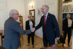 Mężczyzna w garniturze podaje rękę innemu mężczyźnie, w tle kobiety w formalnych strojach.