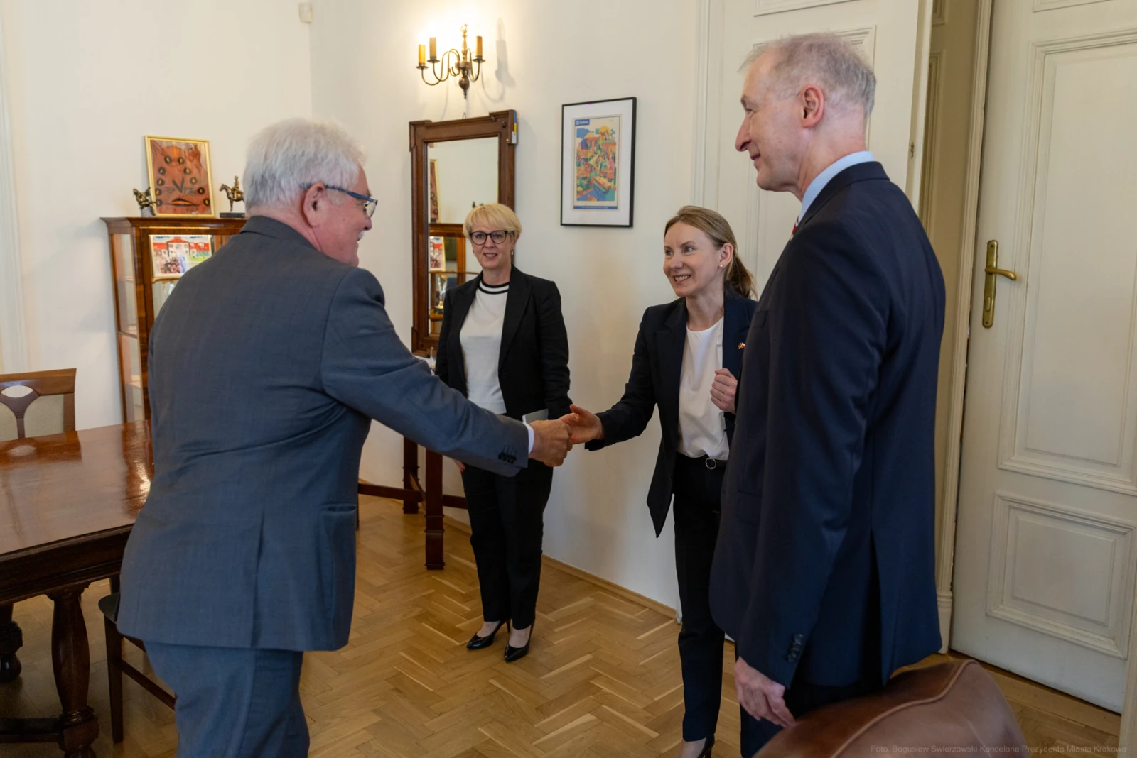 Spotkanie dwóch osób przy uścisku dłoni, w tle kobieta i mężczyzna w formalnych strojach.  Autor: Bogusław Świerzowski