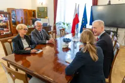 Spotkanie czterech osób w sali konferencyjnej z flagami Polski i UE w tle.