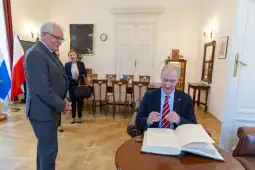 Mężczyzna w garniturze podpisuje księgę, obok stoi inny mężczyzna i kobieta.