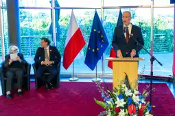 Mężczyzna przemawia przy mównicy, obok siedzą dwie osoby, w tle flagi Polski i Unii Europejskiej.