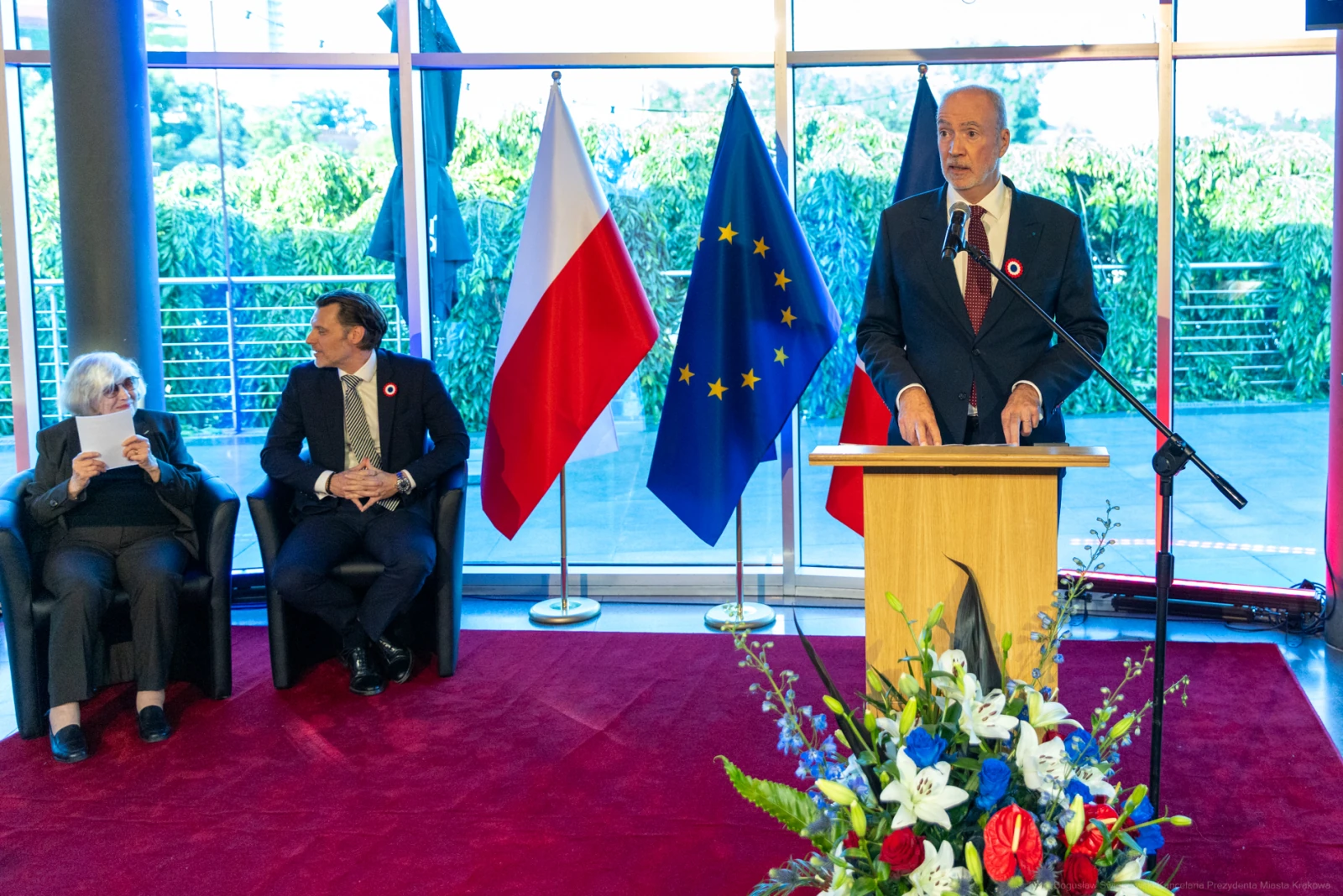 Mężczyzna przemawia przy mównicy, obok siedzą dwie osoby, w tle flagi Polski i Unii Europejskiej.  Autor: Bogusław Świerzowski