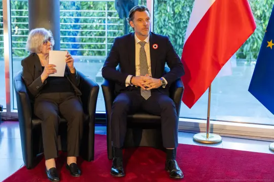 Dwoje ludzi siedzących na kanapie, obok flagi Polski i Unii Europejskiej.