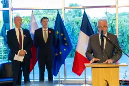 Mężczyzna przemawia przy mównicy, obok dwóch mężczyzn w garniturach, w tle flagi Polski, UE i Francji.