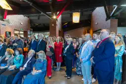 Tłum ludzi w eleganckich strojach, z zainteresowaniem słuchających wystąpienia.