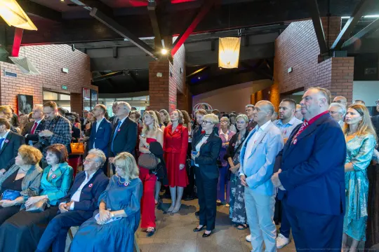 Tłum ludzi w eleganckich strojach, z zainteresowaniem słuchających wystąpienia.