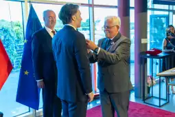 Ceremonia wręczenia odznaczenia mężczyźnie w garniturze. W tle flagi i goście.