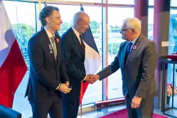 Trzech mężczyzn w garniturach, uściski dłoni, flagi Polski i Francji w tle.